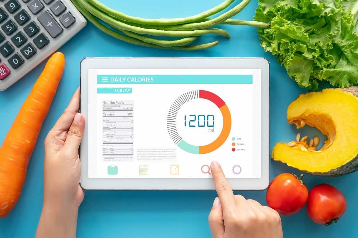 woman using Calorie counter application on tablet at dining table with fresh vegetable and calculator.