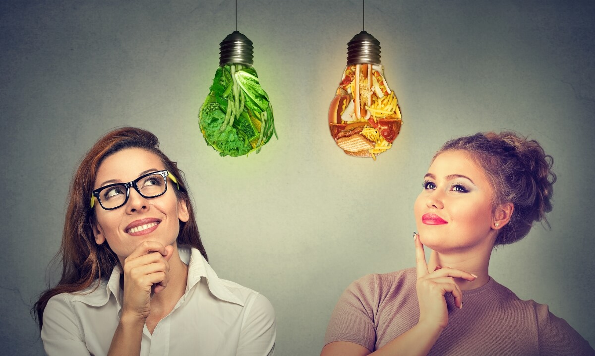Two women thinking about diet one looking at green vegetables light bulb another at junk food lightbulb.