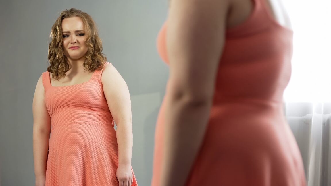 Young female looking in mirror with disgust, ashamed of fat body, obesity issue